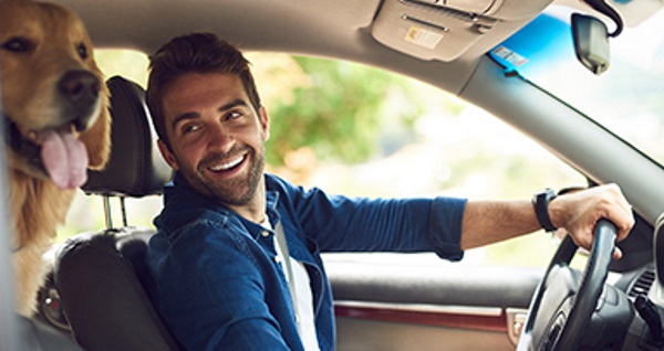 Un homme souriant conduisant une voiture avec un chien sur le siège passager.